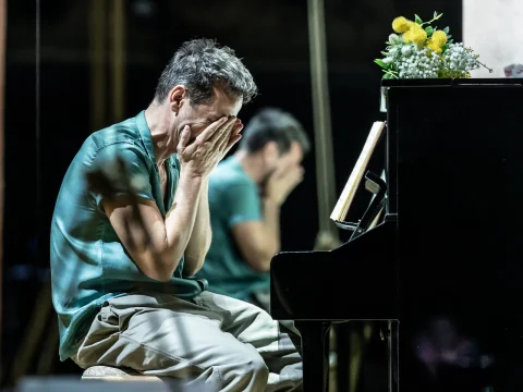 A man sits at a piano with his face in his hands, appearing distressed. A vase with flowers is on the piano. His reflection is seen in a mirror behind him.