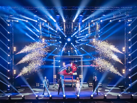 Four performers stand on stage with bright spotlights, fireworks, and a red helicopter prop behind them in a dramatic, theatrical setting.