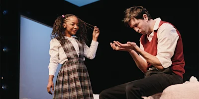 A young girl in a plaid school uniform stands smiling, while an adult man sitting nearby looks down at his hands with a serious expression.