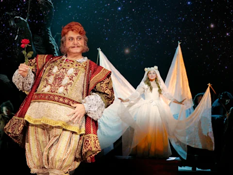 A man in ornate Renaissance-style costume holds a rose, while a woman in a flowing white gown stands with outstretched arms on a stage with a starry backdrop.