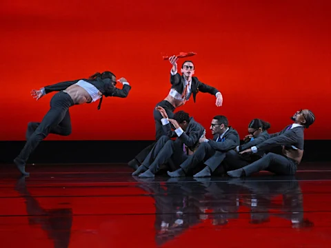 Six dancers in suits perform on stage; one leaps left, while another stands and holds a red object up. The background is a solid red color.