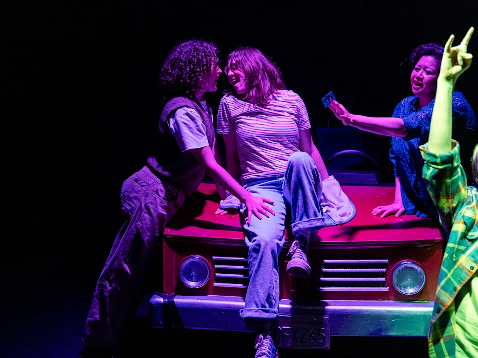 Three people interact animatedly on and around the hood of a red car under stage lighting, with a fourth person’s arm visible in the foreground.