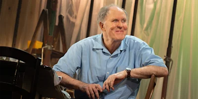 An older man in a light blue short-sleeve shirt sits and smiles in a warmly lit room with wooden furniture and draped fabric in the background.