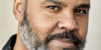 Close-up of a middle-aged man with a salt-and-pepper beard, mustache, and short hair, looking directly at the camera with a neutral expression.