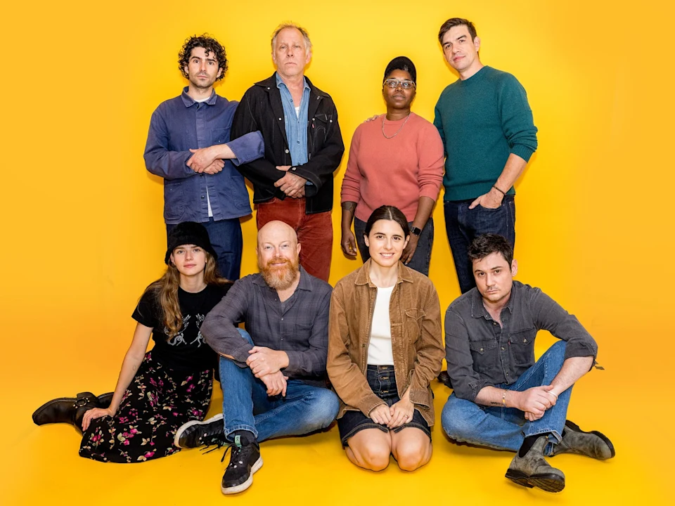 Eight people pose together in two rows against a bright yellow background, with four standing at the back and four seated or kneeling in front.