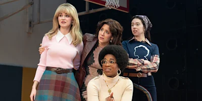 Four women stand together indoors, looking with concern at something off camera; one is seated, and the setting appears to be a gymnasium.