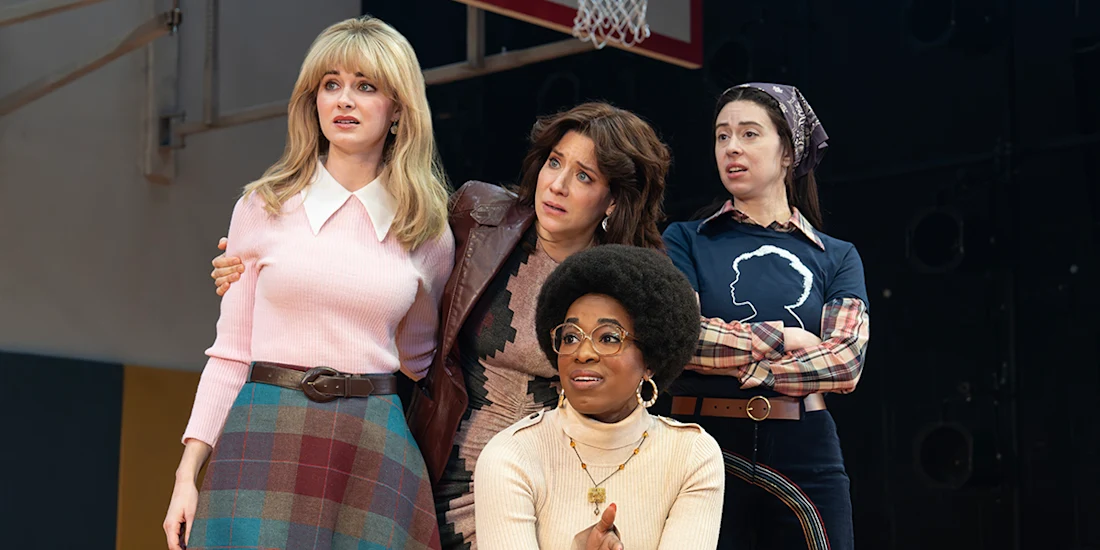 Four women stand together indoors, looking with concern at something off camera; one is seated, and the setting appears to be a gymnasium.