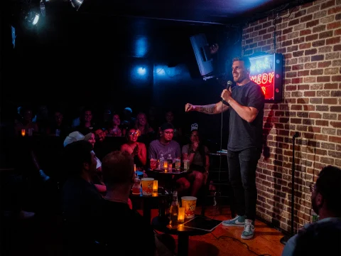 A comedian performs on stage with a microphone in hand, facing an audience in a dimly lit comedy club. A neon sign on a brick wall reads "Comedy." People are seated in the foreground, watching.