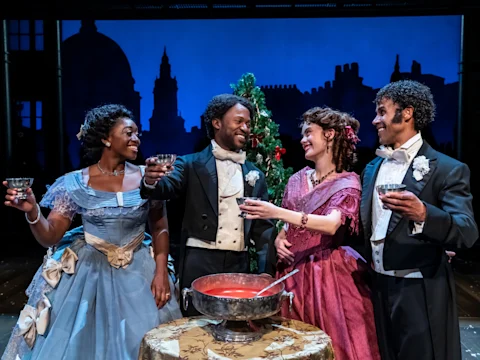 Four people in formal 19th-century attire smile and raise drinks around a table with a punch bowl, with festive decorations and a blue cityscape backdrop behind them.