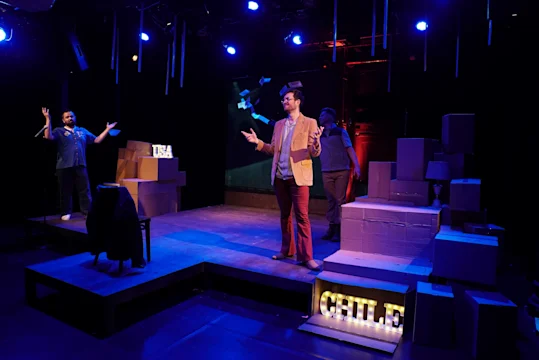 Three actors perform on a stage with cardboard boxes and illuminated letters spelling "CITILE." Blue and purple stage lights highlight the scene.
