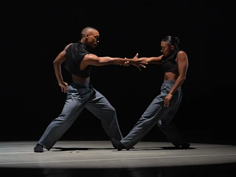 Two dancers in black tops and gray pants perform a contemporary routine on stage, with one extending a hand to the other. The background is dark, highlighting their movements.