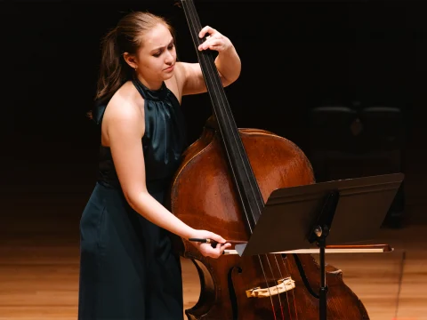 A person in a green dress plays a double bass on stage, reading sheet music from a stand.