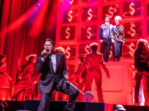 A performer on stage sings into a microphone amid dramatic red lighting, with two people standing on a raised platform in the background surrounded by dollar sign symbols.