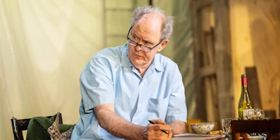 An older man in a light blue shirt sits at a table with papers, a pen, a bottle of wine, and a bowl, looking to the side with a serious expression.