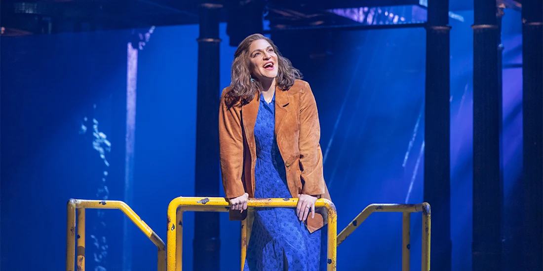 A woman in a blue dress and brown jacket stands behind a yellow railing on a stage with blue lighting and columns in the background.