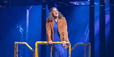 A woman in a blue dress and brown jacket stands behind a yellow railing on a stage with blue lighting and columns in the background.