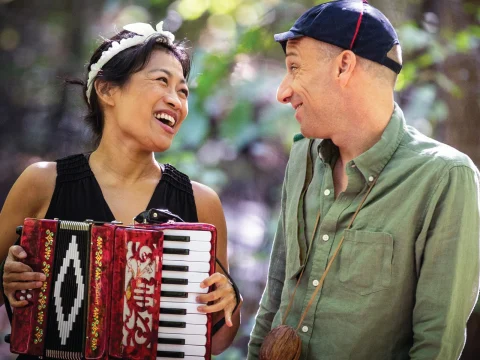 Two people smiling at each other outdoors; one is holding an accordion, and they are surrounded by blurred greenery.