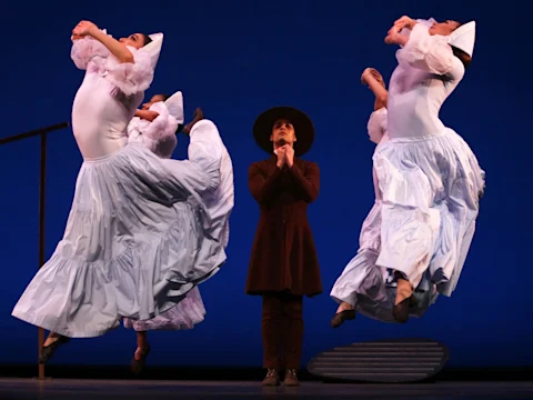 Three dancers in white costumes leap with arms raised on a stage, while a person in a dark outfit and wide-brimmed hat stands with hands clasped at center.