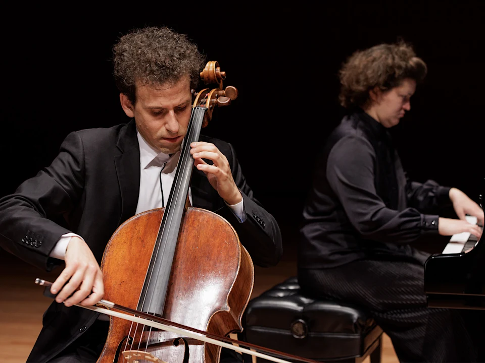 A man in a suit plays a cello with a bow while a woman in dark clothing plays a grand piano in the background on stage.