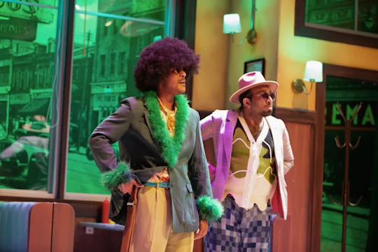 Two men in colorful 1970s-style outfits stand on a stage set resembling a diner. One wears sunglasses and a hat; both have flamboyant clothes and retro hairstyles.