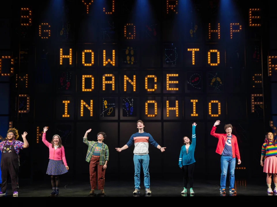 Production photo of How To Dance In Ohio Broadway Reunion Concert in New York City, showing a group of six actors on stage with colorful lighting and digital screens showing symbols and text in the background.