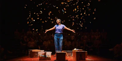 A person stands center stage with arms outstretched, surrounded by falling confetti, with an audience seated in the background and various boxes on the floor.