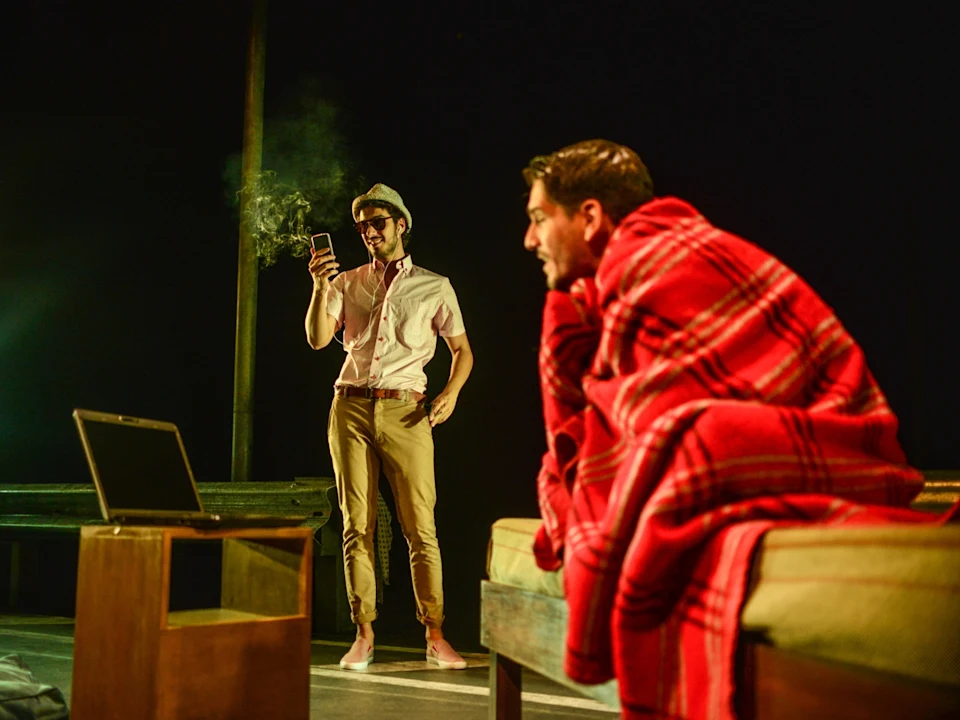 Two men on stage; one stands smiling and looking at a phone, the other sits on a bed wrapped in a red plaid blanket, with a laptop and small table nearby.