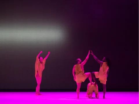 Four dancers in beige costumes perform a contemporary dance routine on stage, under purple lighting. One dancer stands on another's back while two others strike poses with raised arms.