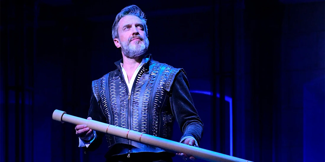 A man in a patterned jacket stands on stage under blue lighting, holding a long, cylindrical object and looking upward.