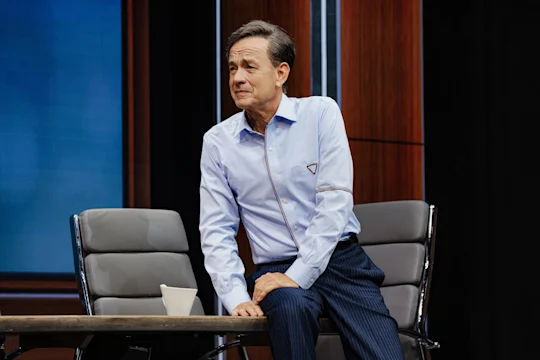 A man in a light blue shirt and dark striped pants leans on a desk beside a chair and a white cup in a modern office setting.