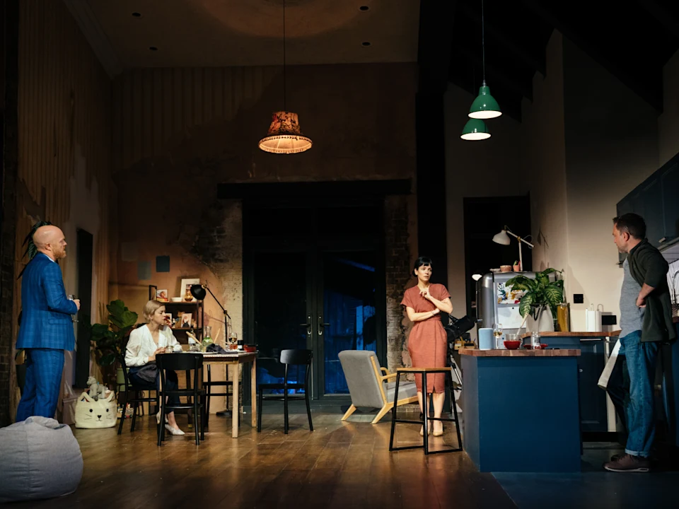 Four adults are standing and talking in a stylish, dimly lit living room and kitchen space with modern furniture and warm lighting.