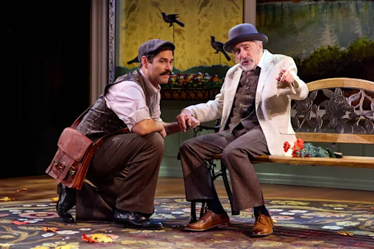 Two men in period costumes talk on stage; one is kneeling with a satchel, the other sits on a decorative bench, gesturing with his hand. Autumn leaves are scattered on the floor.