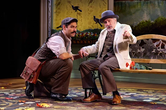 Two men in period costumes talk on stage; one is kneeling with a satchel, the other sits on a decorative bench, gesturing with his hand. Autumn leaves are scattered on the floor.
