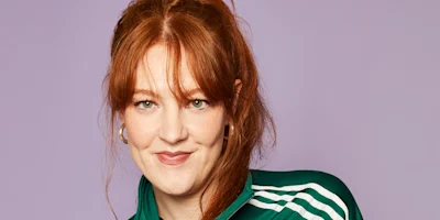 A person with long red hair and hoop earrings wears a green jacket with white stripes, standing against a plain light purple background.