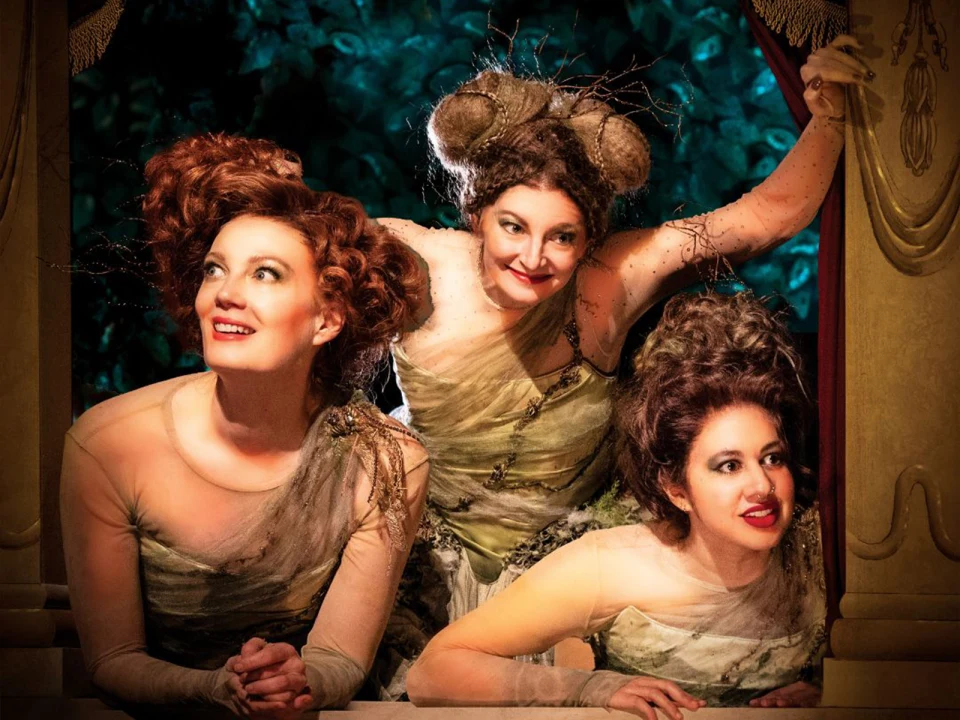 Three women with elaborate hairstyles and costumes resembling forest elements, pose in a theatrical setting with a dark leafy backdrop. Two lean on a window ledge while one stands behind them holding a curtain.