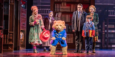 Actors on stage with a person in a Paddington Bear costume surrounded by four adults and a child, set against a backdrop of bookshelves.