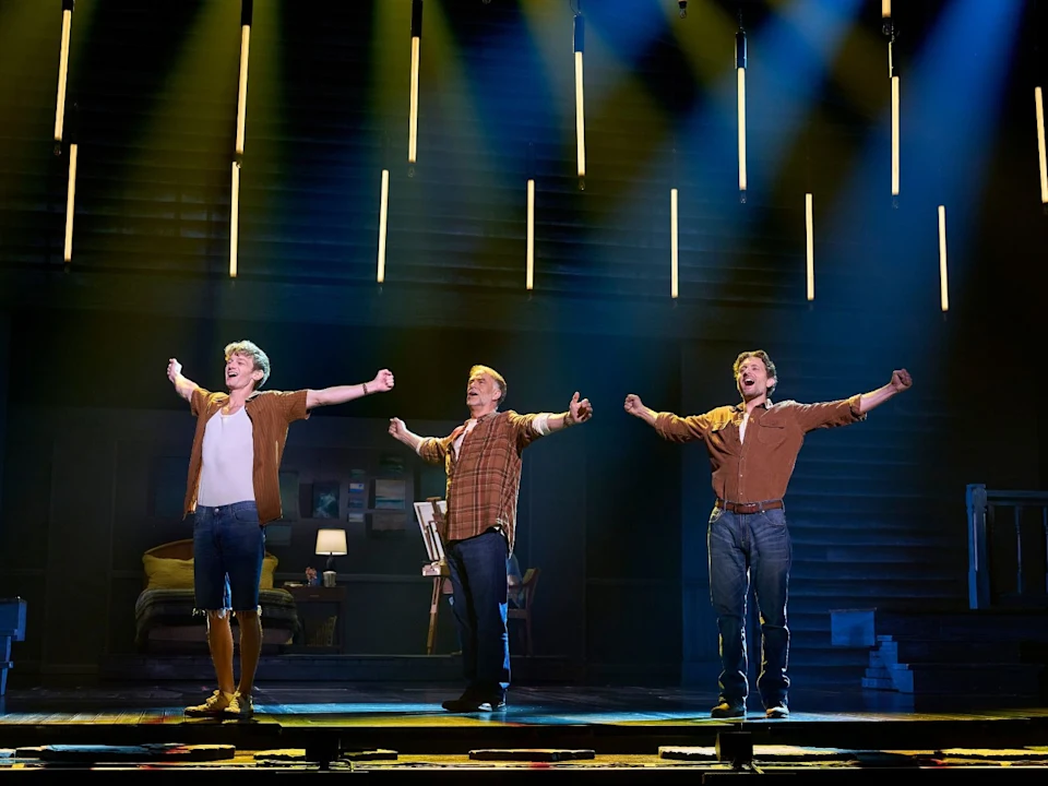 Production photo of The Notebook in Costa Mesa, showing three actors stand on stage with arms outstretched, under vertical lights, performing in a set designed to look like a room with furniture and stairs.