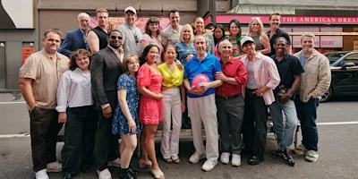 A diverse group of people, both men and women, pose together for a photo on a city street, with some standing and others kneeling, all smiling at the camera.