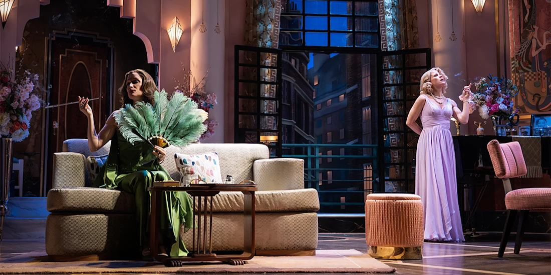 Two women in elegant dresses pose in a lavish living room set; one sits on a sofa with a fan, the other stands by a window holding a cigarette.