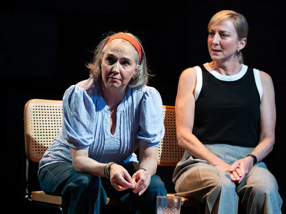 Two women sitting on a chair, one wearing a light blue blouse and headband, the other in a black sleeveless top and gray pants, both looking attentively in the same direction.