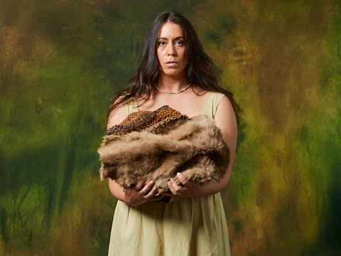 A woman in a beige dress stands against a mottled green and brown background, holding a bundle of animal fur and patterned fabric.