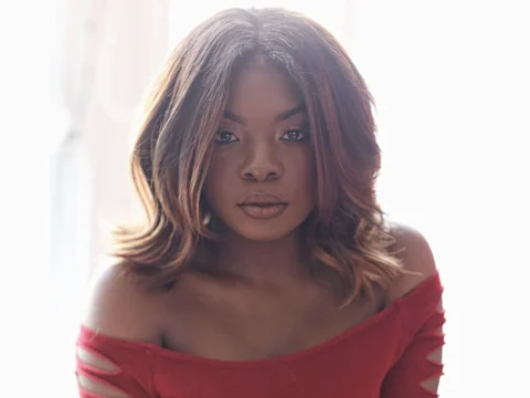 Woman with shoulder-length wavy hair wearing a red off-the-shoulder top, looking directly at the camera with a neutral expression.