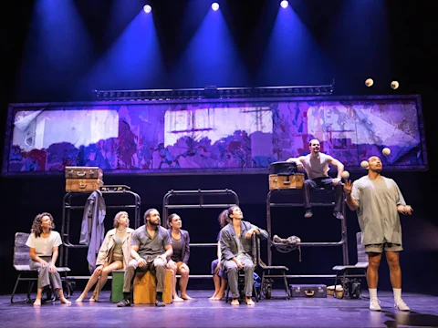 Seven people sit on or near bunk beds on stage under blue lights, while one person stands to the right juggling three balls. Some suitcases are placed nearby.