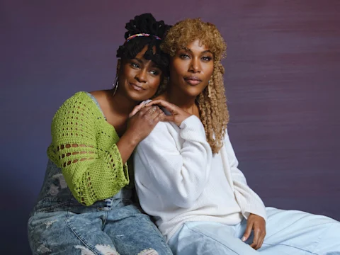 Two people sit closely on a bench. One wears a green sweater and overalls, the other a white sweater. They both have curly hair and are holding hands against a purple background.