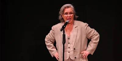 A person with gray hair and glasses speaks into a microphone on stage, wearing a beige suit with hands on hips against a black background.