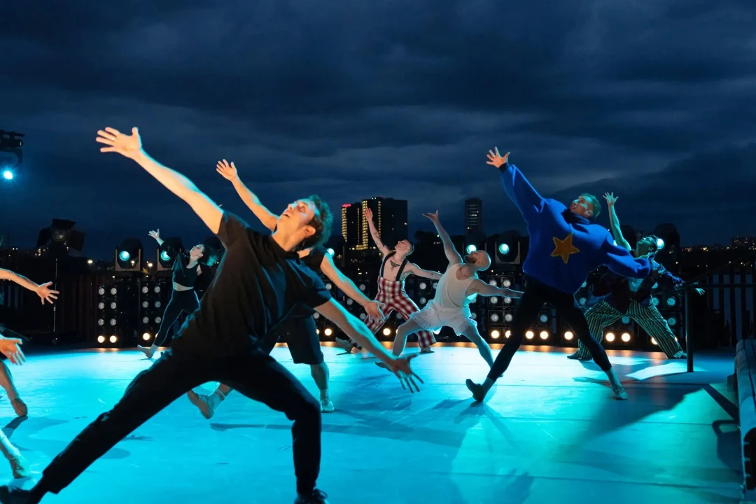 A group of dancers perform a dynamic routine on an outdoor stage at night, with city buildings and a dark, cloudy sky in the background.