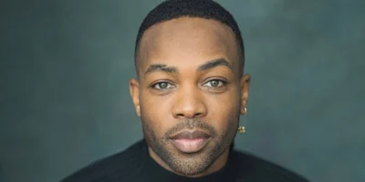 A person with short hair and an earring looks directly at the camera, wearing a black top against a dark, blurred background.