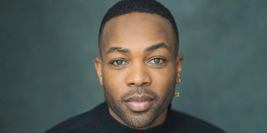 A person with short hair and an earring looks directly at the camera, wearing a black top against a dark, blurred background.