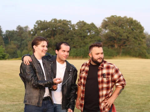 Three men standing outdoors on a grassy field, two wearing leather jackets and one wearing a plaid shirt. They appear to be socializing, with trees in the background.