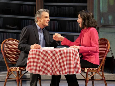 A man and woman sit at a checkered tablecloth-covered table, holding hands and smiling at each other in a room with a bookshelf in the background.